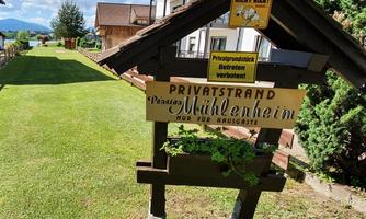 A sign reading "Private Beach Pension Mühlenheim" stands in a well-kept garden. In the background, houses and trees can be seen, as well as a blue sky.