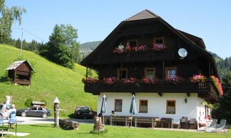 A traditional wooden house with blooming balconies in a green environment. Various cars are parked in front of it, and there are outdoor seating areas.