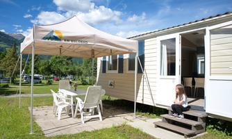A cozy mobile home at a campsite with a sunshade and seating. In the foreground, a girl is sitting on the steps and enjoying nature.