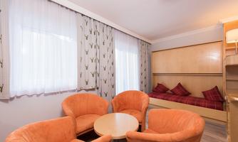 A cozy room with orange chairs and a small table. In the background, there is a bed with red pillows and light curtains.