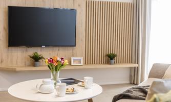 A modern living room with a flat-screen TV on the wall. On a round table, there is a vase of flowers, cups, and snacks.