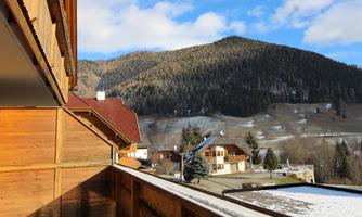 A balcony view of a mountainous landscape with snow-covered hills and traditional wooden houses. The sky is clear and sunny.