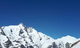 An impressive mountain landscape with snow-capped peaks and a bright blue sky. In the foreground, there is a wooden railing that borders the view.