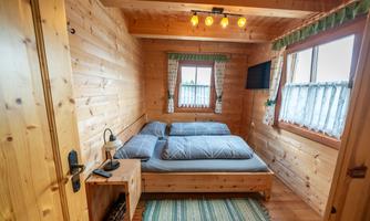 A cozy wooden bedroom with a double bed and a small nightstand. The walls and ceiling are also made of wood, and there is a window with curtains.