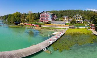 A picturesque lake with a wooden pier and green shore areas. In the background, colorful buildings and trees can be seen.