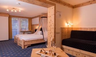 A cozy room with a double bed and a sofa. The room is designed in warm wood tones and has large windows with curtains.