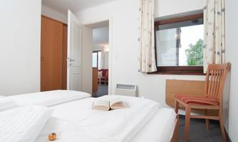 A simple bedroom with two beds, a wooden chair, and a window. A book lies on the bedspread and the room feels bright and inviting.