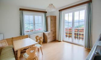 A cozy dining area with a wooden table and chairs. Large windows offer a view of the mountains and the balcony.