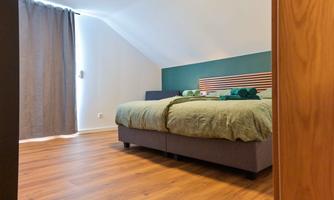 A modern bedroom with a large bed and green bedding. Fresh towels lie next to the bed, and a window with a dark curtain ensures privacy.