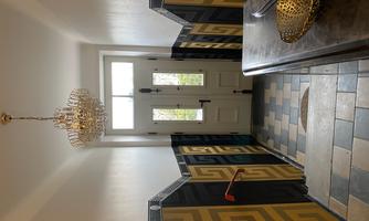 A bright hallway with an elegant ceiling light and ornate walls in yellow and black. The front door is open and lets in lots of light.
