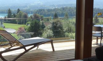 A relaxing view from a balcony overlooking a beautiful lake and green landscapes. A deck chair and a table with chairs invite you to linger.