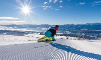 A skier is skiing down a groomed slope in bright sunshine. In the background, mountains and a clear sky can be seen.