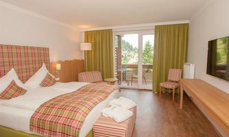 A cozy hotel room with a large bed and red checked pillows. The room has a balcony and large windows overlooking the greenery.