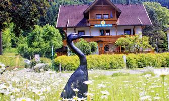 A picturesque inn in the background, surrounded by green trees and meadows. In the foreground stands a black sculpture of a swan amidst flowers.