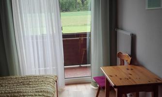 A simple room with a bed, a table, and two chairs. The balcony offers a view of a green landscape.