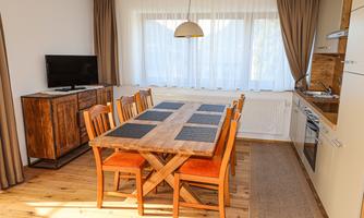 An inviting dining area with a large wooden table and chairs. The open kitchen is bright and friendly, with a window that lets in plenty of daylight.