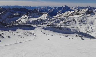 An impressive mountain landscape with snow-covered slopes and a clear blue sky. In the distance, more mountains and skiers on the slope can be seen.