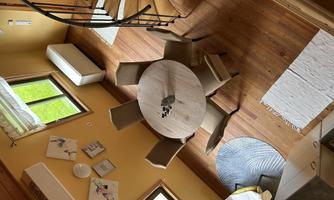 A cozy dining area with a round table and six chairs. Large windows let in plenty of daylight, and the floor is made of wood.