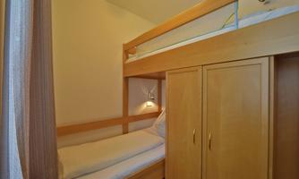 A small room with a wooden bunk bed and a wardrobe. The walls are light and the light is soft.