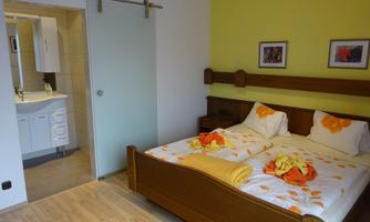 A cozy bedroom with a large bed and colorful bed covers. In the background, you can see a modern bathroom through a glass door.