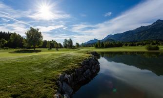 A picturesque golf course with gentle hills and trees. In the background, the Karawanken are reflected in the clear water of a pond.