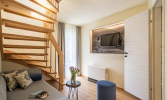 A modern living room with a comfortable couch and a small table. A wooden staircase leads upstairs, and a TV is mounted on the wall.