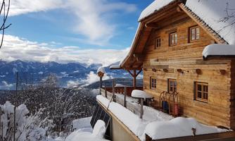 A cozy wooden house in a snowy landscape. In the background, majestic mountains and a blue sky can be seen.