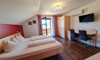 A modern bedroom with a large bed, desk, and chairs. The room has windows facing outside and is bright and inviting.