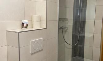 A modern bathroom with a shower and a toilet. The walls are light tiled and there is toilet paper and towels nearby.