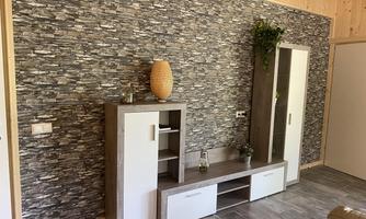 A modern living room with a stone wall and light wood. There is a TV furniture piece decorated with a vase and plants.