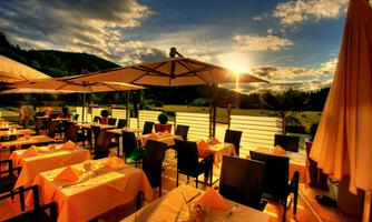A cozy terrace of a restaurant with orange tablecloths and sun umbrellas. In the background, a beautiful sunset and a green landscape are visible.