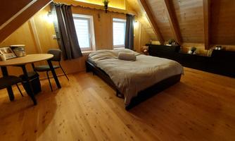 A cozy bedroom with wooden paneling and large windows. The room features a bed, a table, and wooden chairs.
