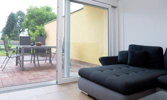 A modern living room with a grey couch and a large window front. Outside is a terrace with a table and chairs as well as lots of green garden.