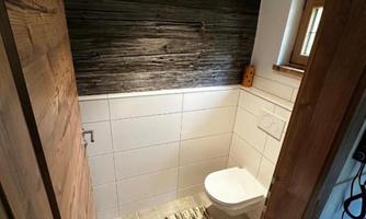 A modern bathroom with a white toilet and a textured wood look on the wall. The floor is covered in gray tiles and a rug is placed in front of the toilet.