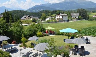 A picturesque landscape with a green garden and various terrace furniture. In the background, mountains and some houses can be seen.