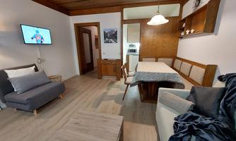 A cozy living room with a gray sofa and a dining area. A television hangs on the wall and there is wood paneling.