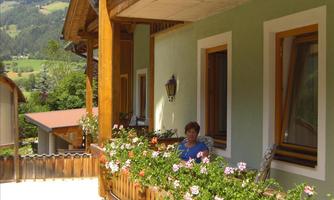 A woman is sitting on a balcony, surrounded by blooming plants. In the background, the mountains and a green landscape can be seen.