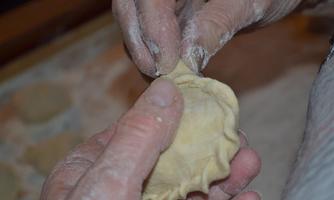 A person is shaping dough for filled dumplings. The hands are dusted with flour, and in the background, more dumplings can be seen.