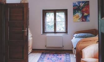 A cozy room with wooden flooring and a colorful carpet. There is a window with a view outside and simple furniture.
