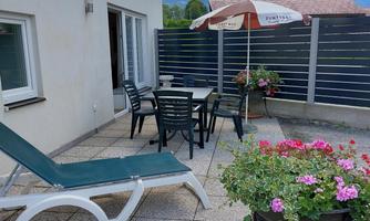 A cozy terrace with a table, chairs, and a lounge chair. In the background, there are flowers and a sunshade.