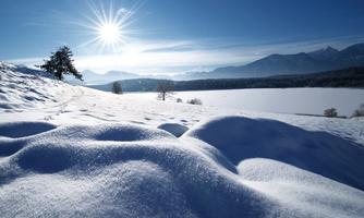 A snow-covered landscape under a blue sky. The sun shines over the mountains in the background.