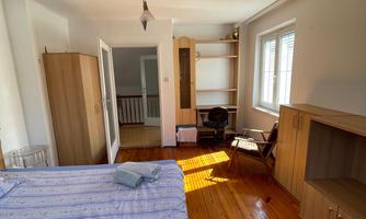 A bright bedroom with wooden flooring and large windows. There is a bed, a desk, and cabinets for storage.
