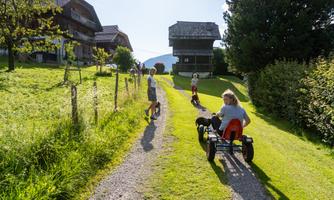A group of children is playing on a path in nature. In the background, traditional houses and plenty of green grass can be seen.
