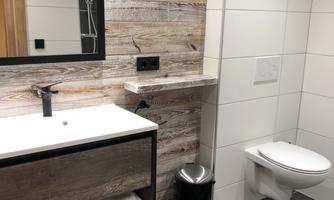 A modern bathroom with a wooden shelf, a sink, and a wall mirror. The floor is tiled in dark stone look, and there is a modern toilet area.
