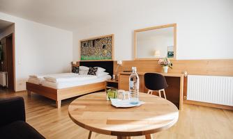 A modern bedroom with a large bed, a table, and chairs. The walls are light and simple, with some decorative elements.