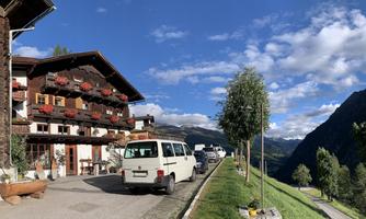 A traditional alpine house with blooming window boxes and a picturesque view of the mountains. Several vehicles are parked along the road in a quiet, green landscape.