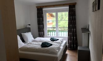 A cozy bedroom with two single beds and a balcony. The walls are light, and large windows offer a view of nature.