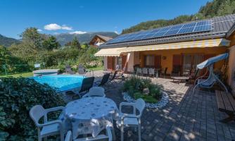 A cozy outdoor area with a pool, sun loungers, and a dining table. In the background, green mountains and a clear sky can be seen.