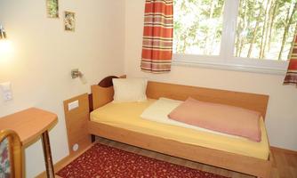 A simple room with a bed, a small table, and a window overlooking trees. The walls are light, and there is a carpet on the floor.