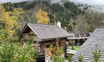 A rustic wooden house surrounded by autumn trees and mountainous landscape. The clouds hang low over the mountains, giving the scene a mystical atmosphere.